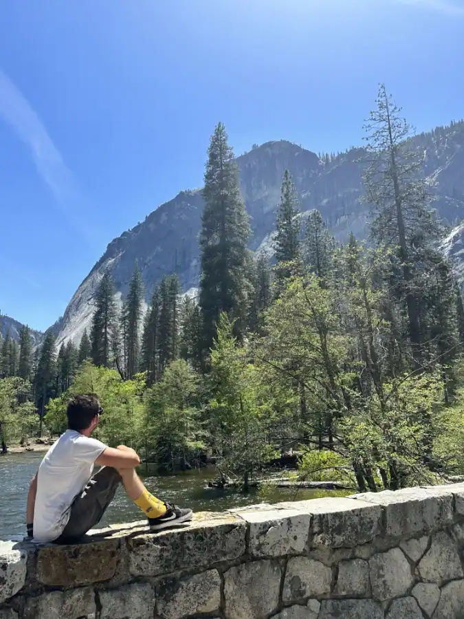 Dean sitting in his wheelchair on a stone wall by a Yosemite bridge with mountains, trees and water behind him